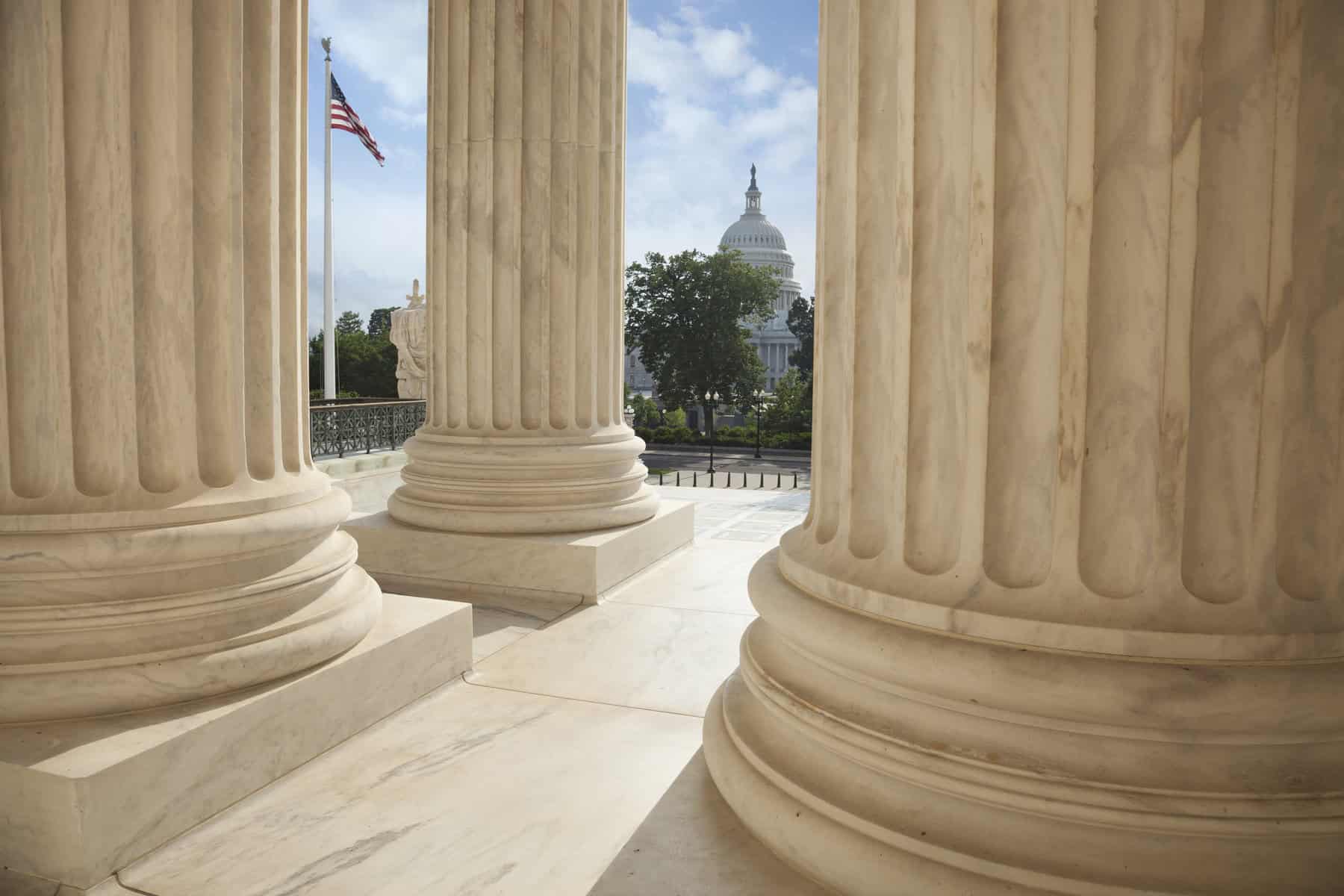 Close up of the columns  of the Supreme Court building with an American flag and the US Capitol in the background